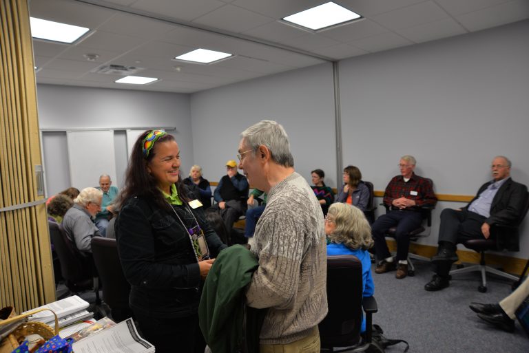 A man and a woman stamp in a conference room chatting with one another. A dozen other adults can be seen in the background, sitting in chairs.