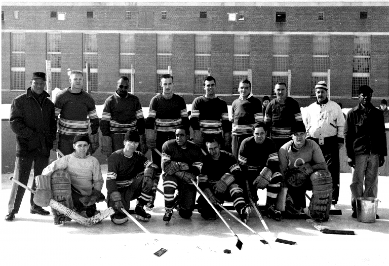 15 men, most of whom are wearing striped hockey jerseys, stand in two rows and pose with their hockey sticks for a group photo