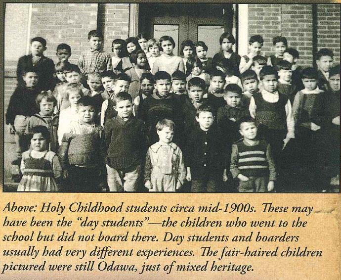 Black and white photo of approximately 40 young students posing for a group photo on the steps of a brick building. Embedded caption reads: "These may have been the 'day students' - the children who went to the school but did not board there. Day students and boarders usually had very different experiences. The fair-haired children pictured were still Odawa, just of mixed heritage."